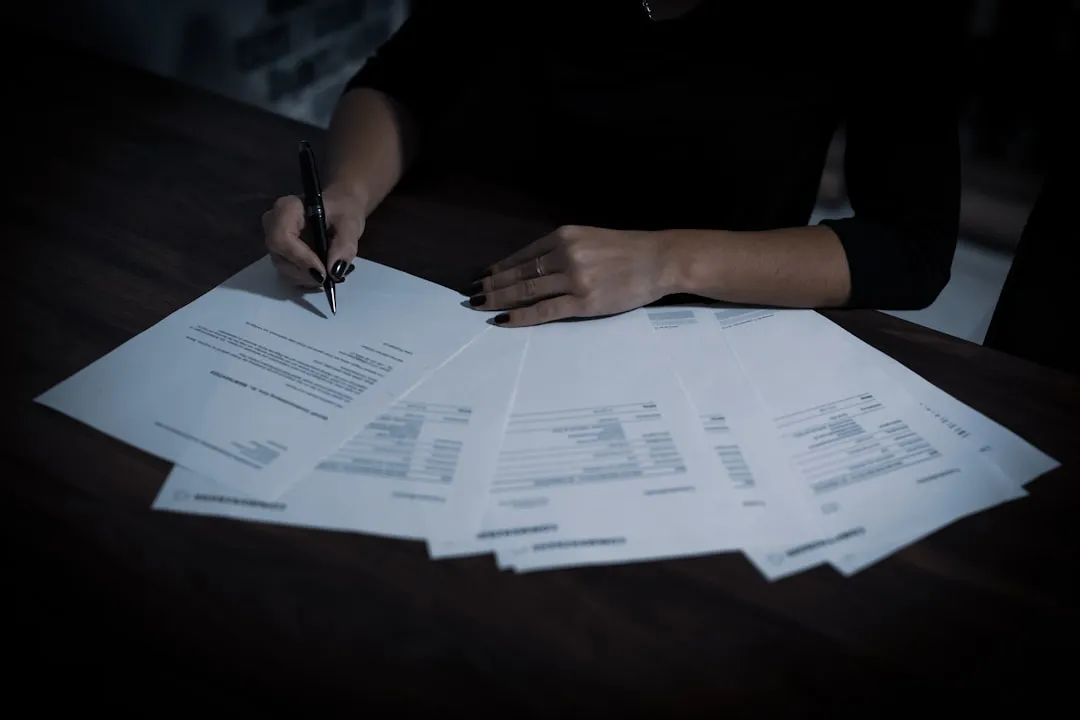 Pessoa organizando documentos em pasta com carimbo e impressora ao fundo, ambiente administrativo.